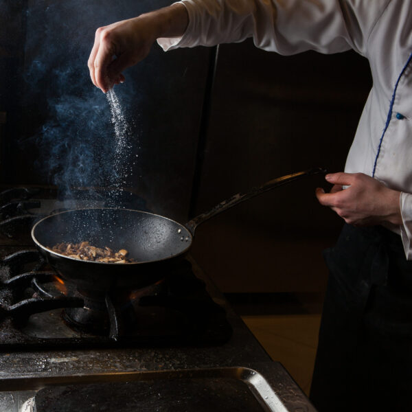 Side view mushroom frying with salt and fire and human hand in pan on black background